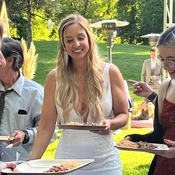 Smiling woman in a white dress holding a plate of Mexican rice and beans at an outdoor event.