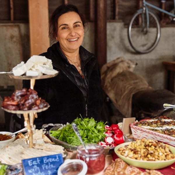 Smiling woman behind a table laden with Middle Eastern food, including feta, dates, and flatbread.