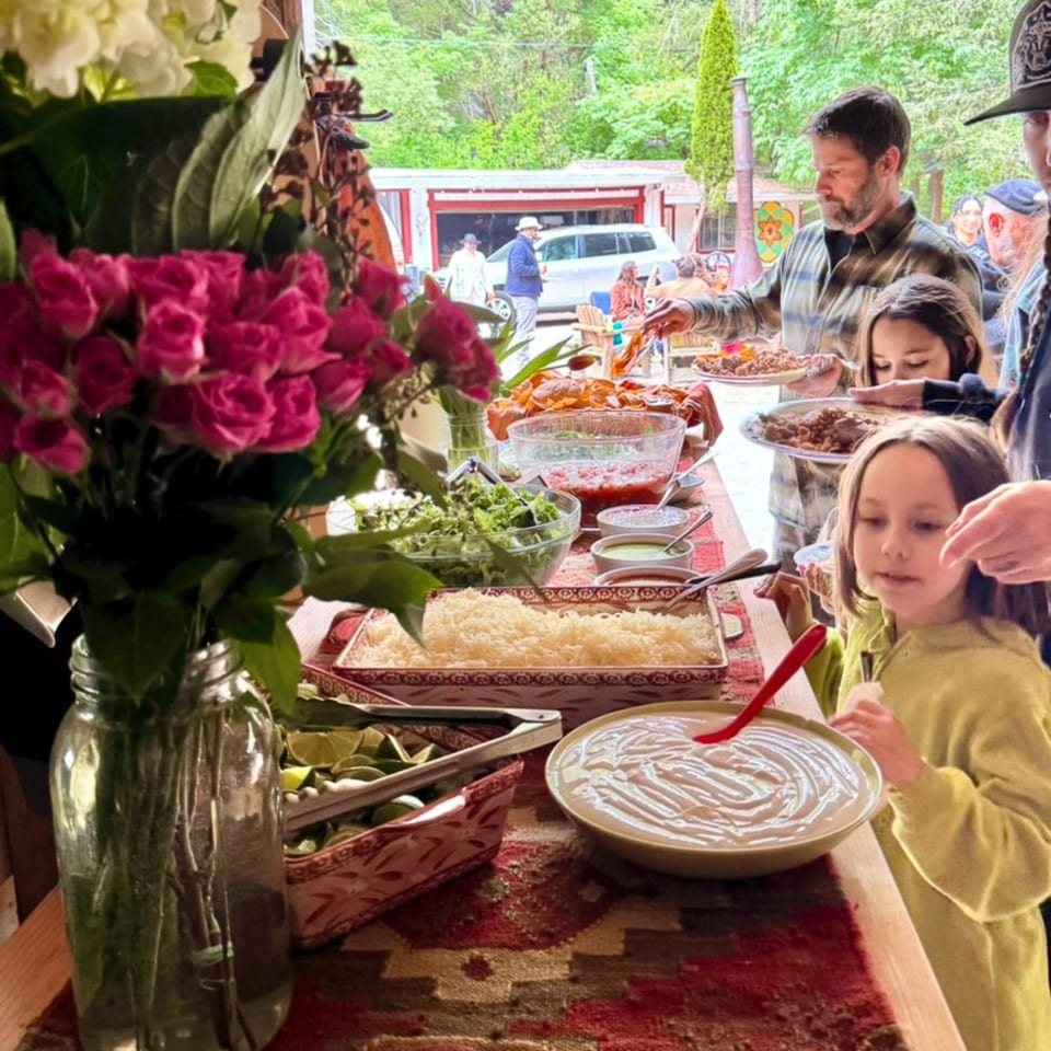 People serving themselves from a buffet table laden with food and flowers at an outdoor gathering.