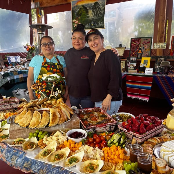 Three women smiling behind a large buffet table laden with various Mexican dishes and snacks.